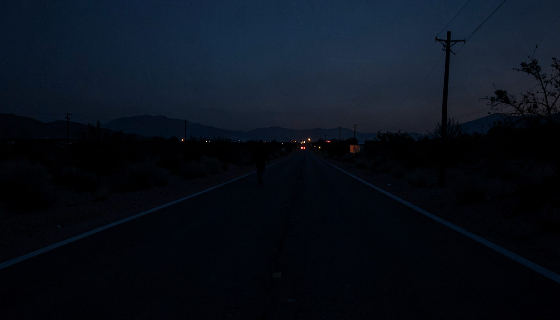 A dim, shadowy desert road at dusk near the US-Mexico border, deep navy and charcoal tones, a lone figure's silhouette walking toward a distant horizon
