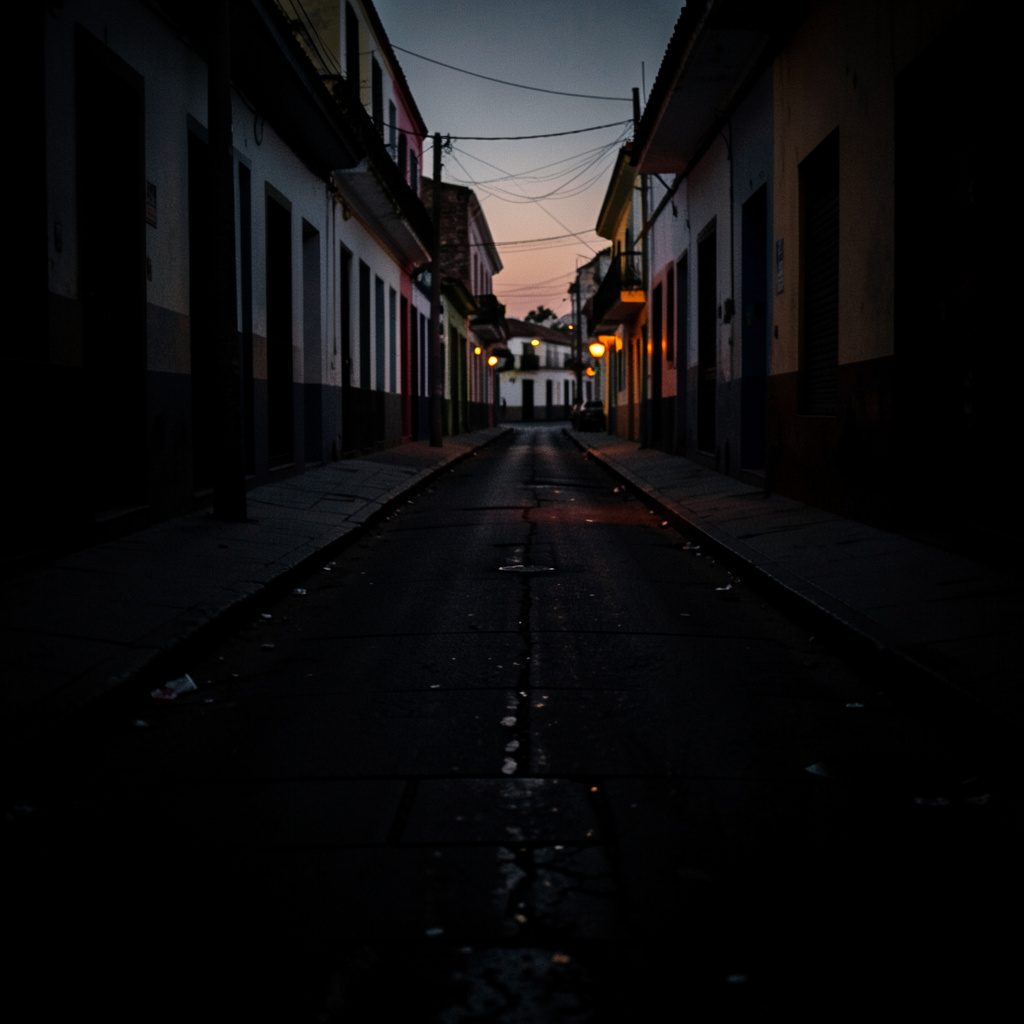A dark, low-key photograph of a quiet street at dusk in a Mexican city, with deep shadows stretching across worn pavement and a subdued, contemplative atmosphere.