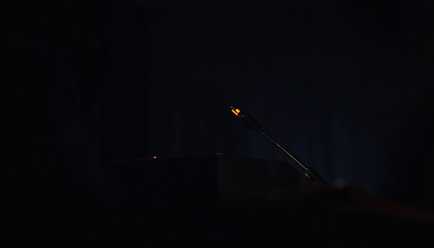 A dark, low-key photograph of a press conference microphone on a lectern, deep shadows across the foreground, muted navy tones