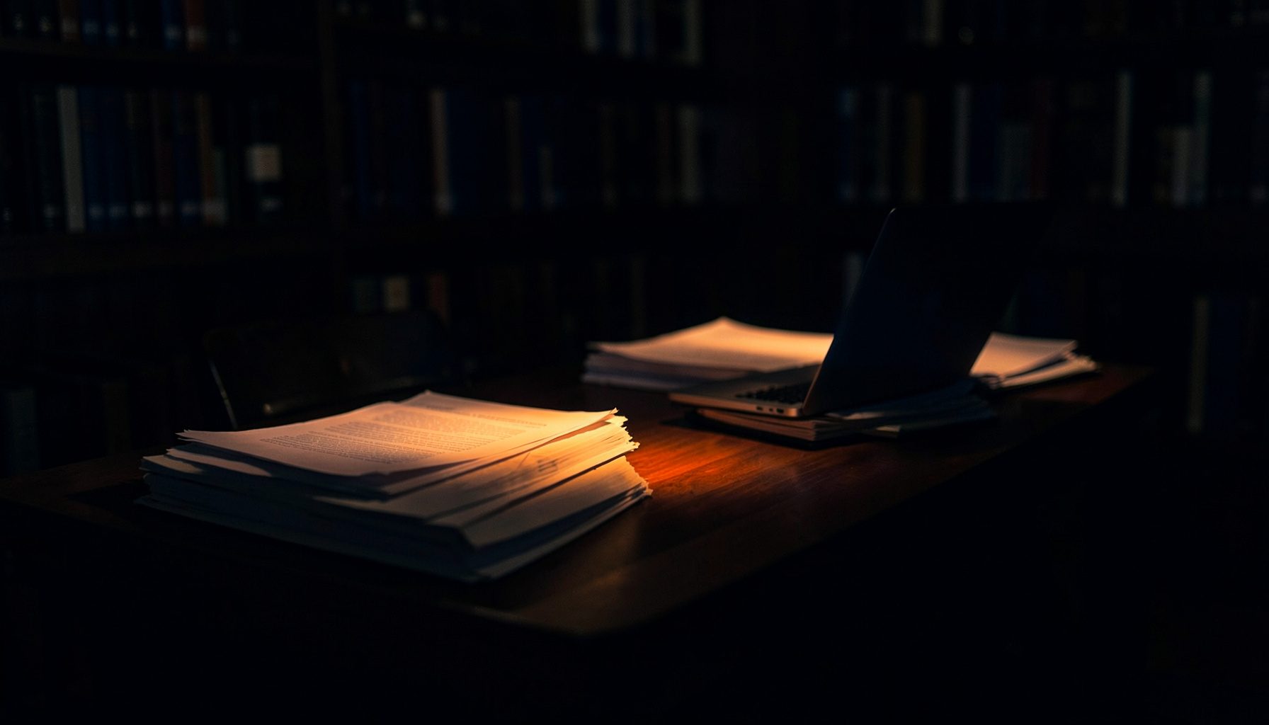 A dimly lit library table with stacked papers and a glowing laptop, deep navy shadows and warm amber highlights across the surface