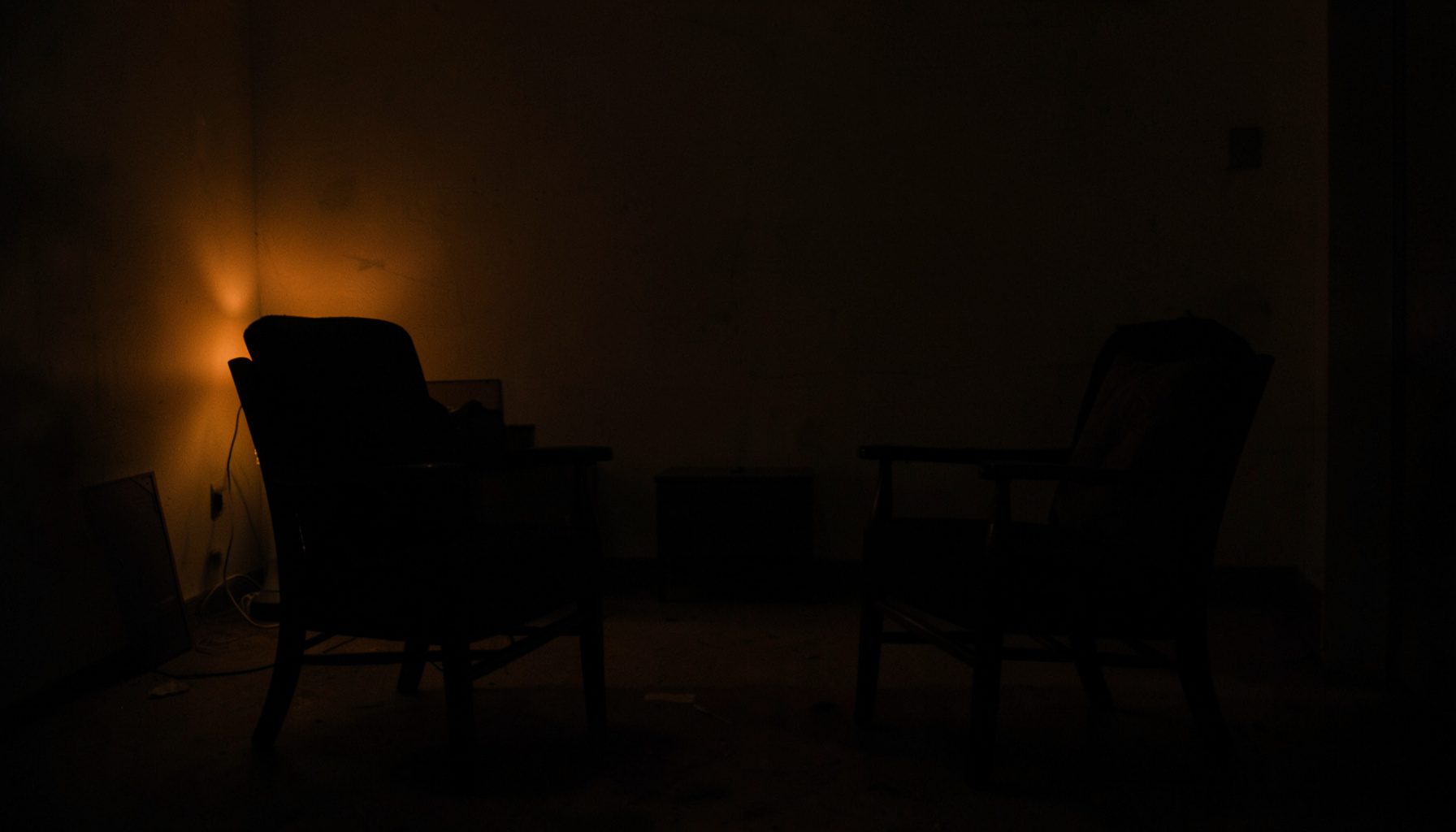 A dark, low-key photograph of two chairs facing each other in a sparsely furnished room, lit by warm lamplight from one side, deep shadows across the floor.