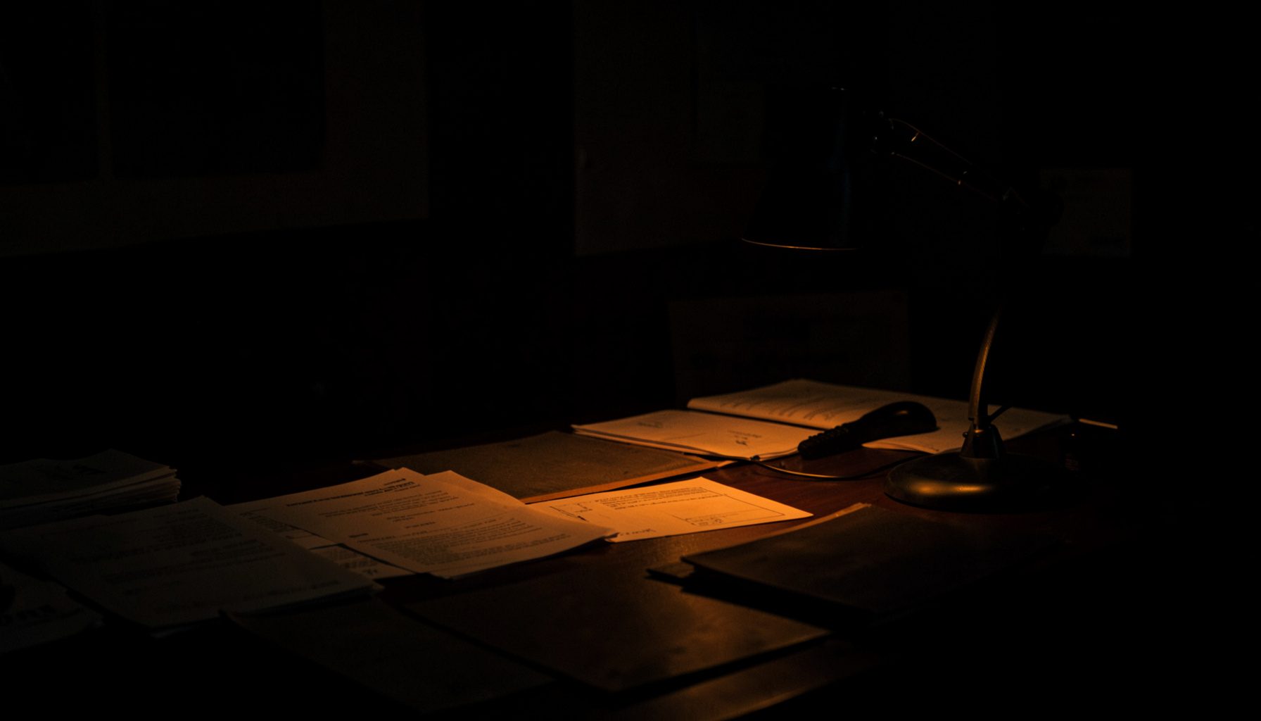 A dark, low-key photograph of a dimly lit newsroom desk at night, scattered papers and a muted desk lamp casting long shadows