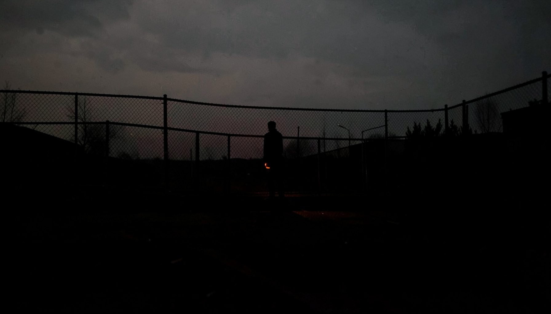 A solitary figure standing at a border fence at dusk, deep shadows stretching across the foreground, evoking the uncertainty and hope of those seeking refuge.