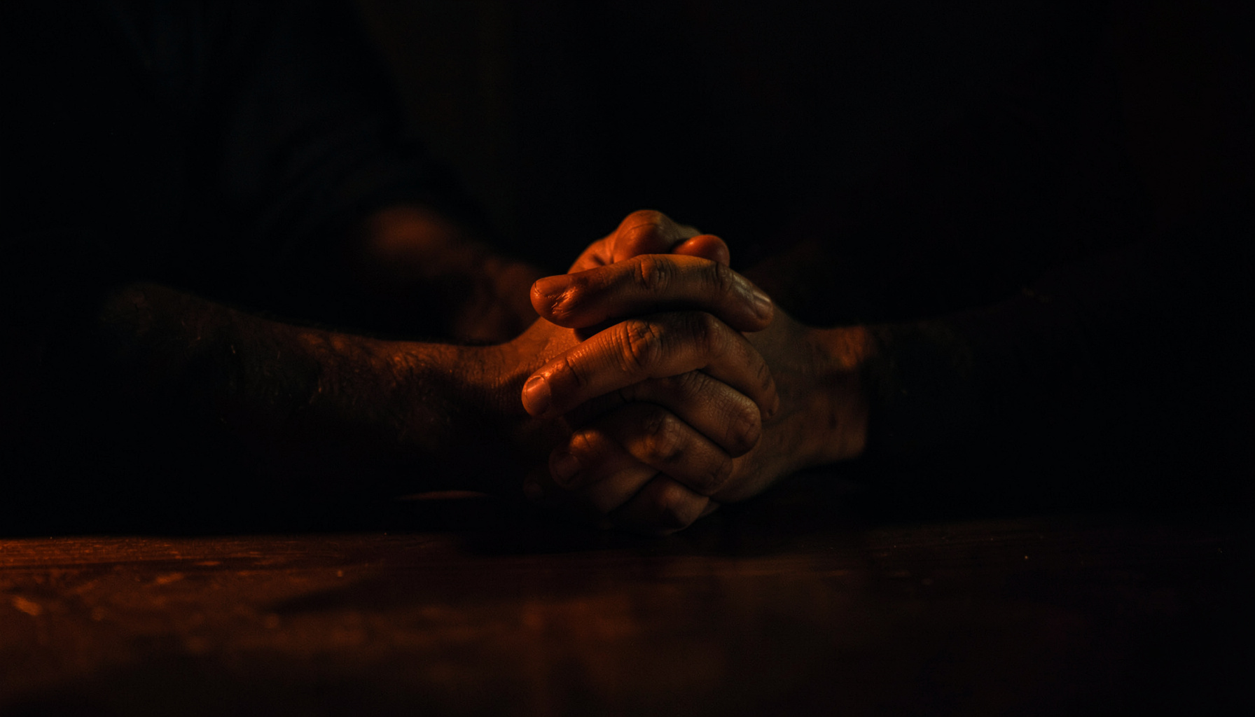Dark, moody photograph of hands clasped together on a table in low light, deep shadow conveying support and quiet determination